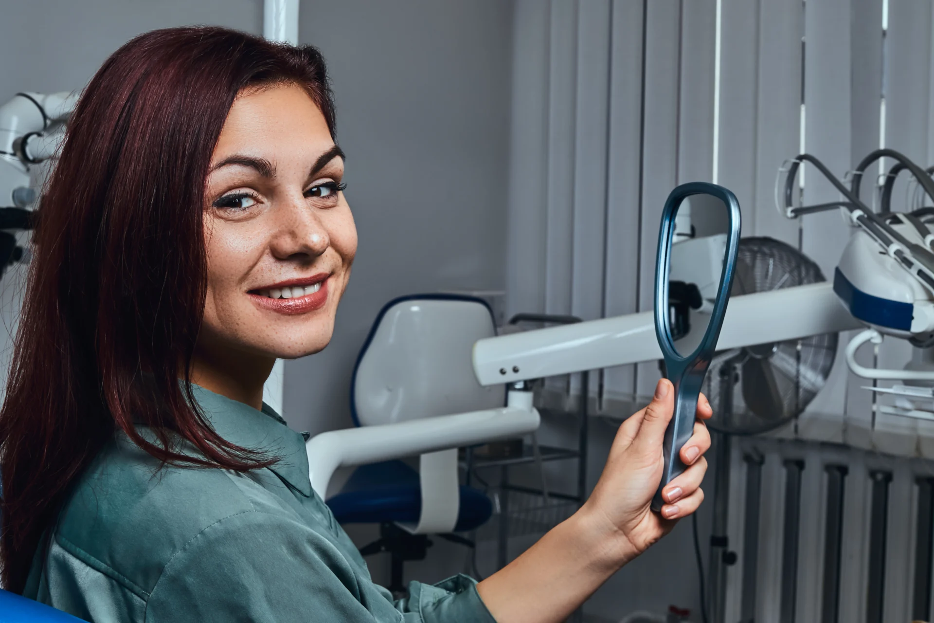 Beautiful smiling redhead girl sitting dentist chair with mirror hands looking camera